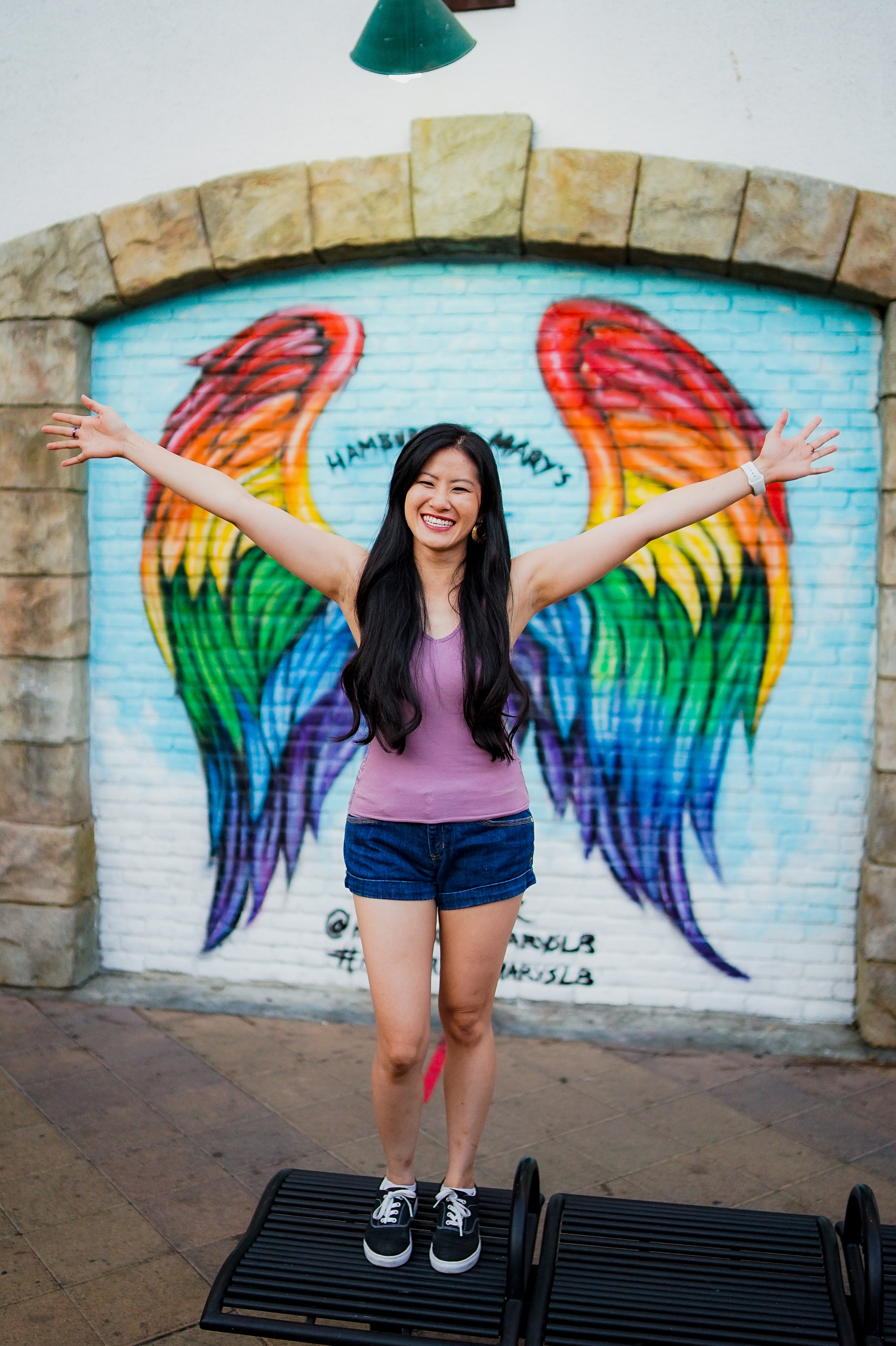Image of a person with long dark slightly wavy hair, a purple tank top, and dark jean shorts. Arms are spread out in front of a wall painting with a blue background and rainbow-colored angel wings