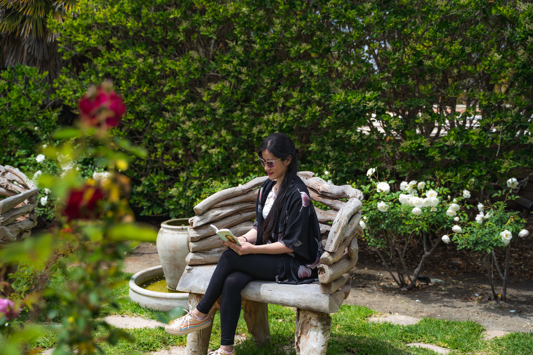 A woman with long dark hair, sunglasses dark clothing, a white top, and pink shoes sitting on a wooden chair reading a book in a garden