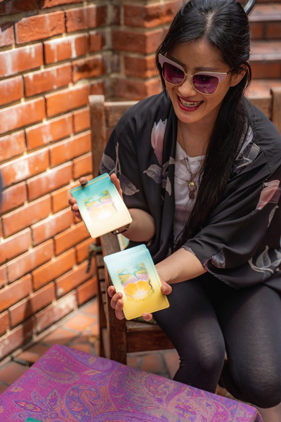 Person smiling with sunglasses and long dark hair shuffling an oracle card deck with a brick wall in the background